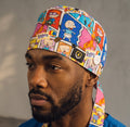 A man wearing a colorful cartoon-print surgical scrub cap and matching scrub ties over blue medical scrubs, photographed against a plain gray wall.