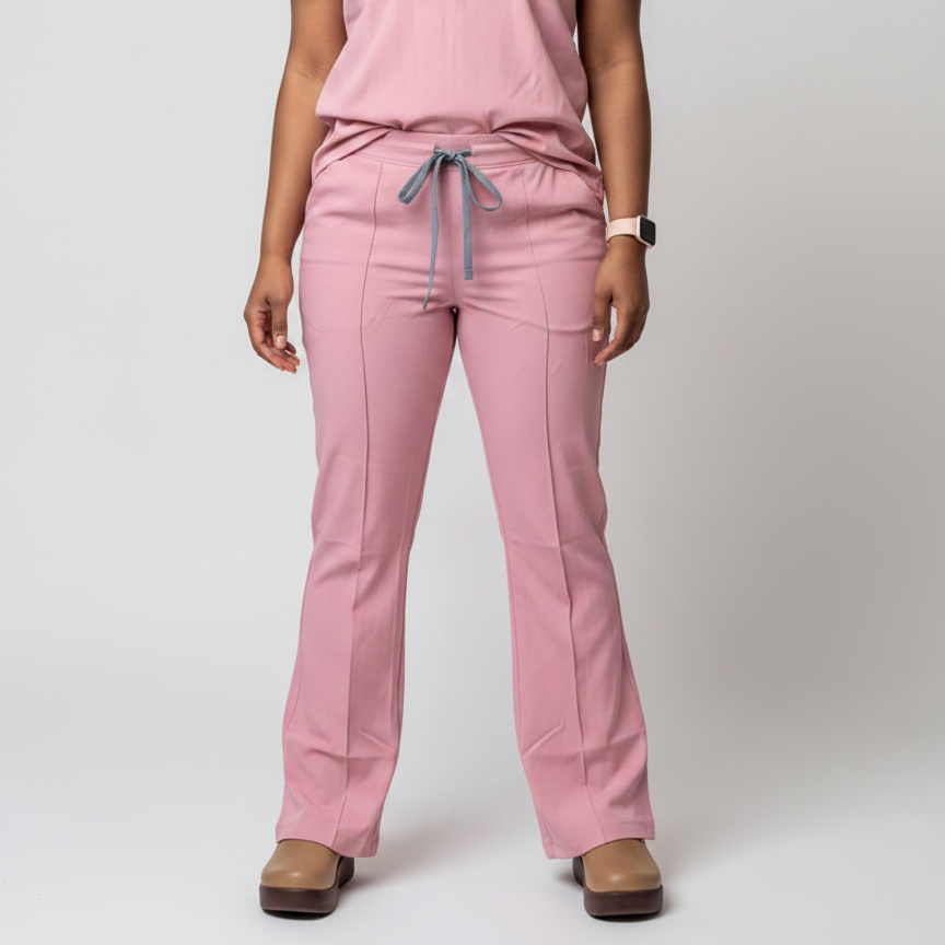 A woman modeling a matching light pink scrub set consisting of a short-sleeve V-neck top and straight-leg pants, standing against a light gray studio background.