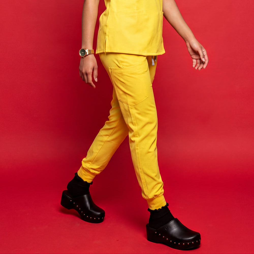 A woman modeling a bright yellow scrub set consisting of a short-sleeve V-neck top and matching pants, paired with black clogs, against a solid red studio background.