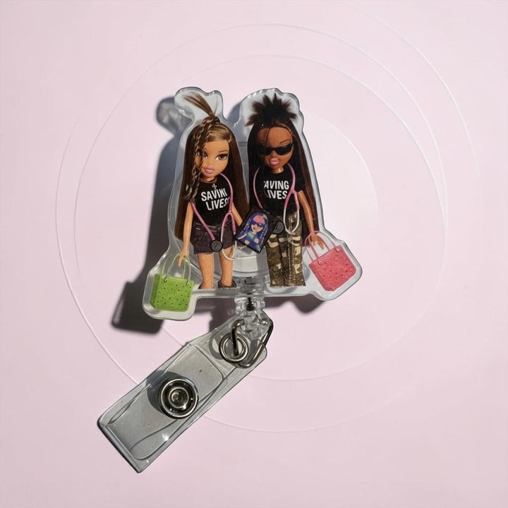 Scrubs Galore Uniforms nurse badge reel with two dolls wearing "Saving Lives" shirts, stethoscopes, and carrying bags.