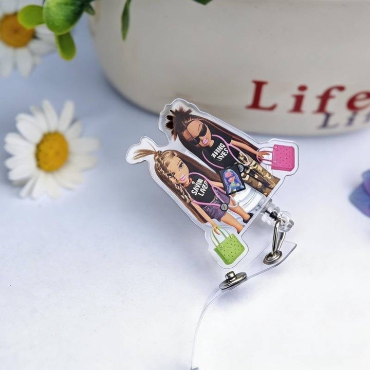 Scrubs Galore Uniforms nurse badge reel featuring two dolls wearing 'Saving Lives' shirts and carrying handbags. Clear acrylic with a retractable clip.